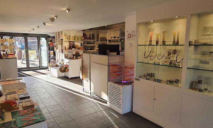 A large glass entrance opens onto tiled floor. Cabinets to the left with cards books and scarfs to the right glass jewelry displays, shelves with bask